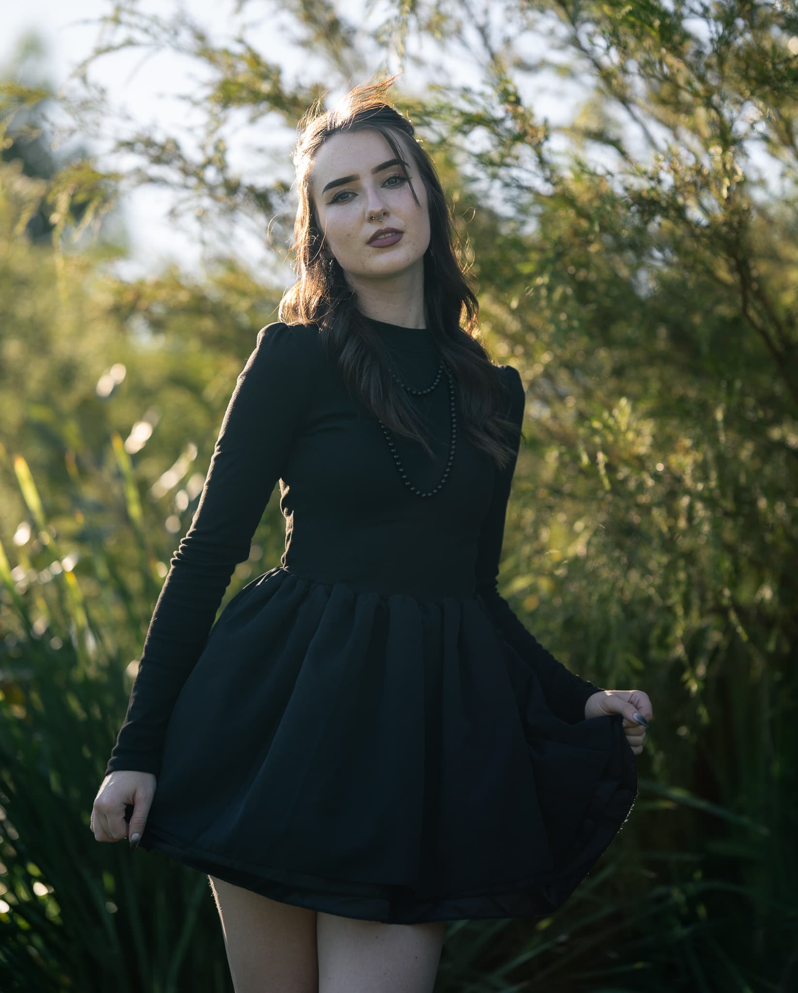 Woman in a black long-sleeve dress holding out the hem of her skirt, backlit by low sun