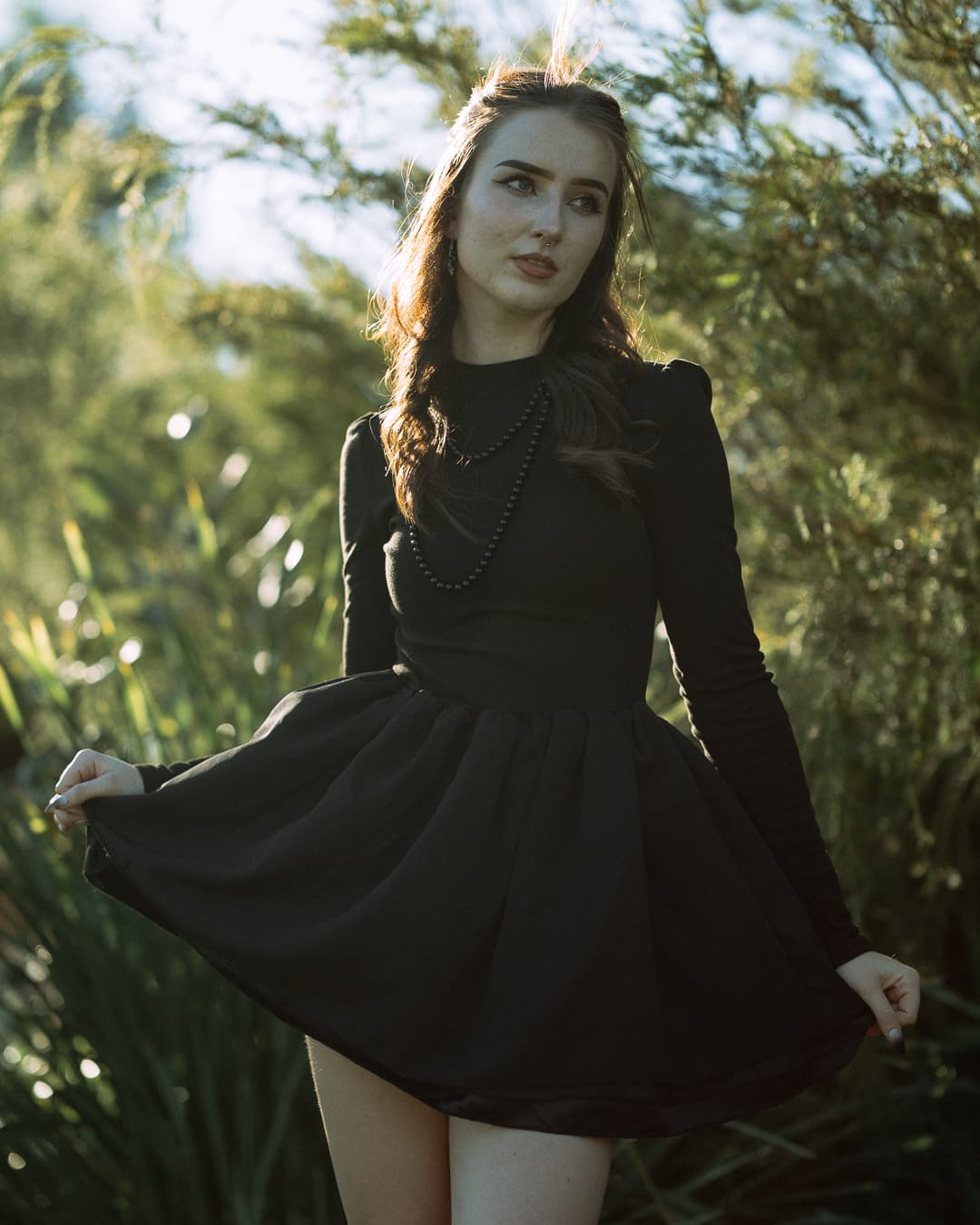 Woman in a black dress and pearl necklace looking away among sunlit grasses