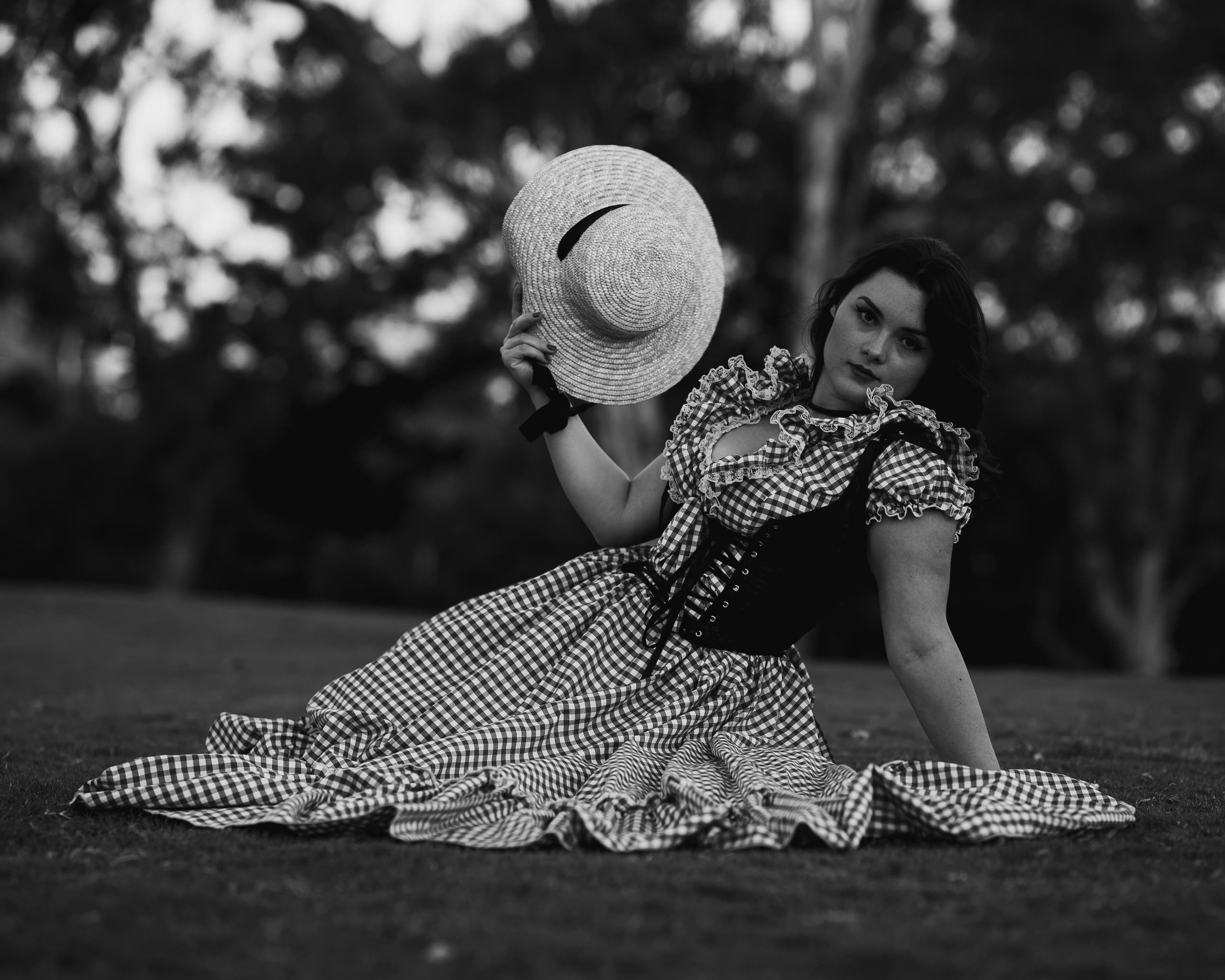 Black and white portrait of a woman in a gingham dress seated on grass, raising a straw hat