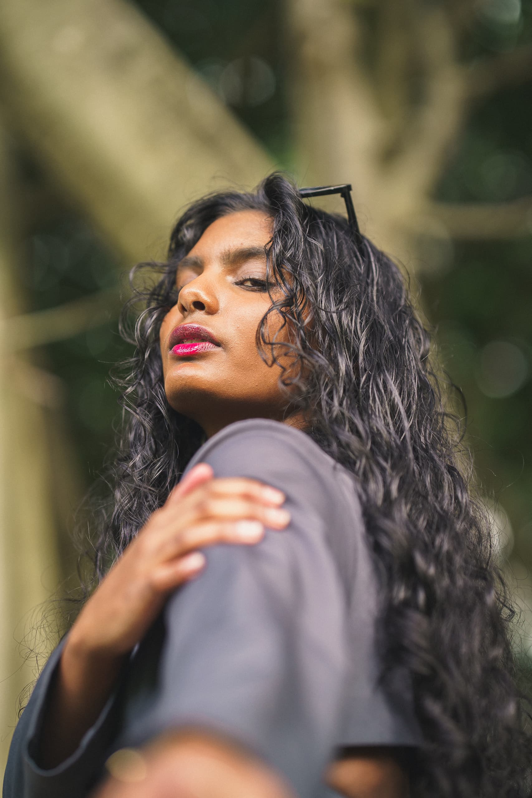 Low-angle portrait of a woman with dark curls and bright pink lips under trees