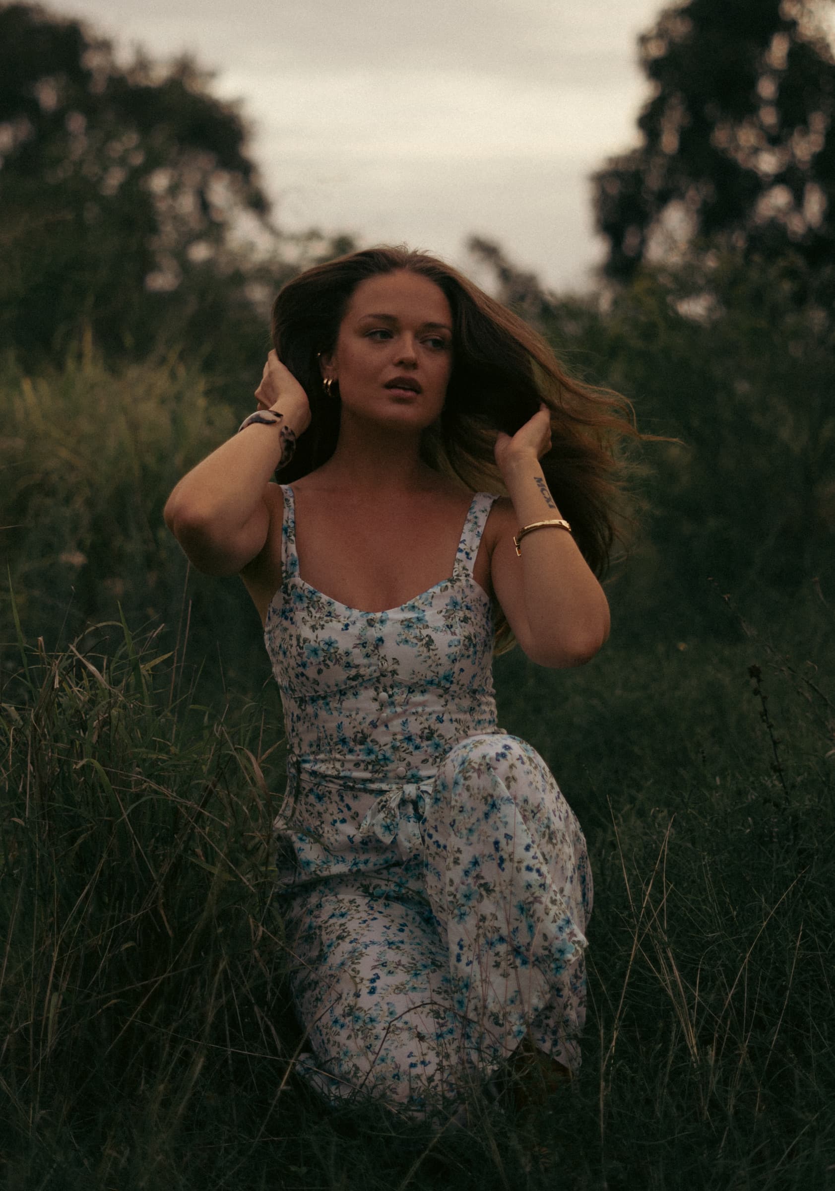 Woman in a floral dress crouched in long grass fixing her hair under an overcast sky