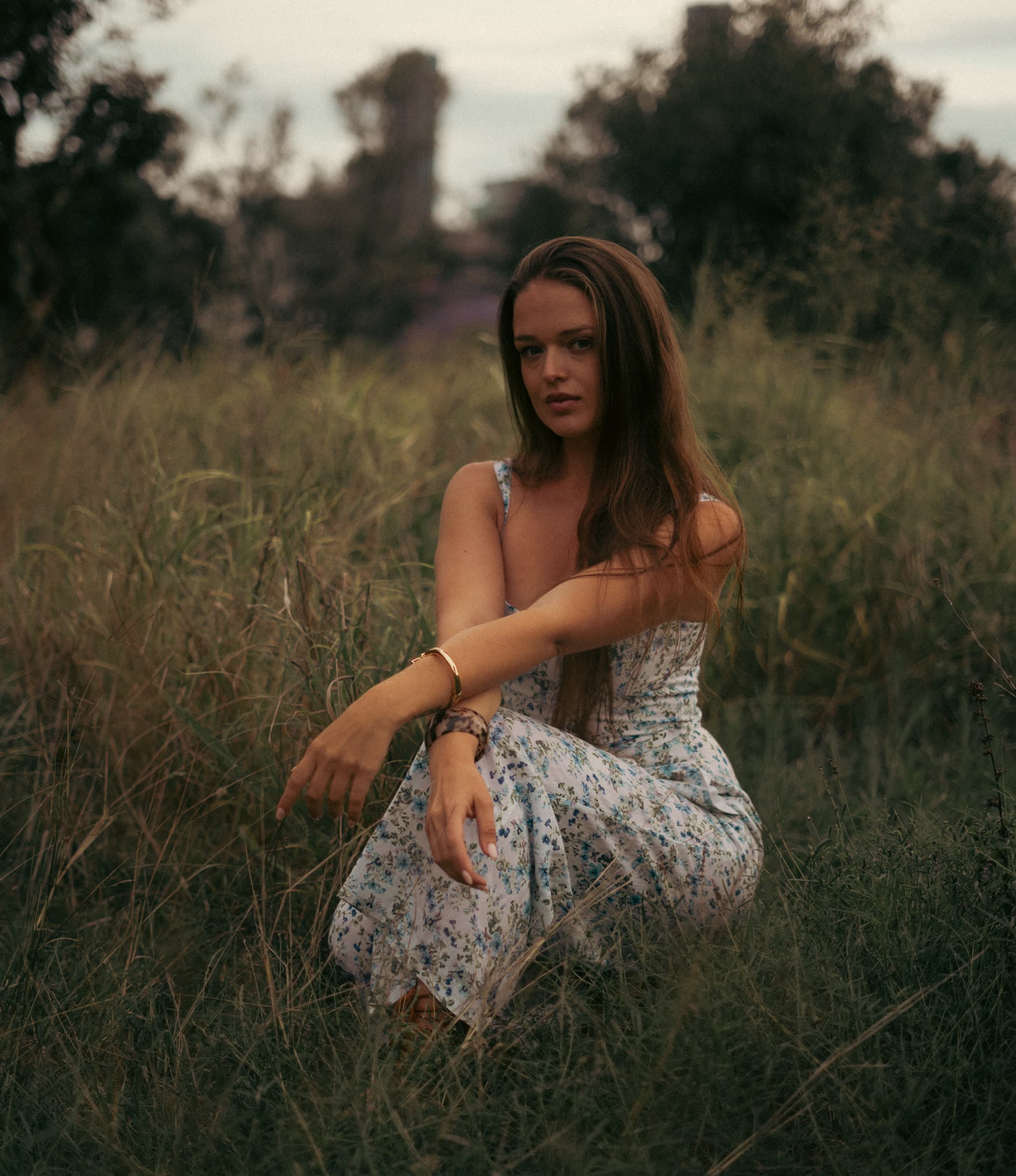Woman in a floral dress crouching in a grassy field, looking straight into the lens