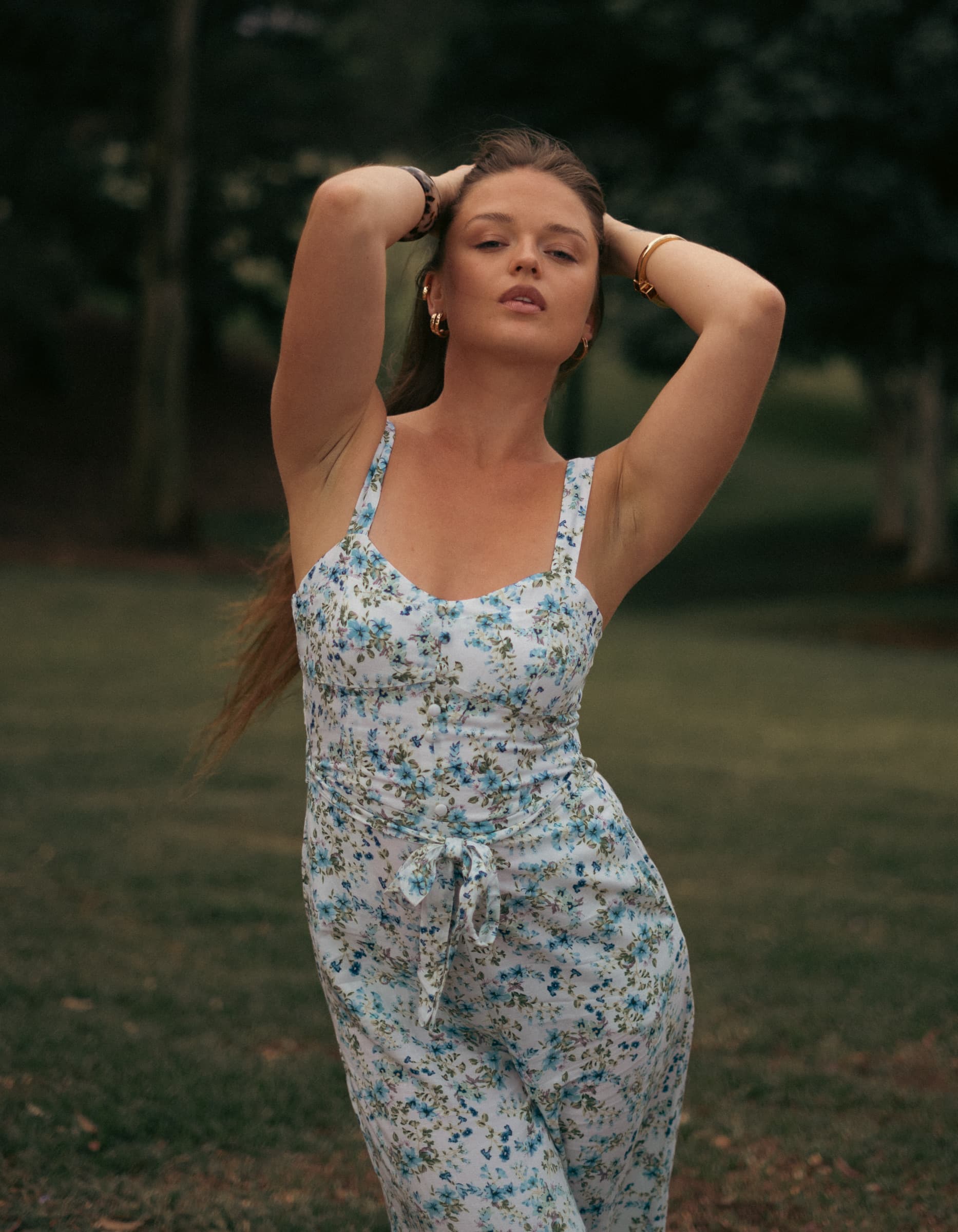 Woman in a floral dress standing in a park at dusk with her arms raised behind her head