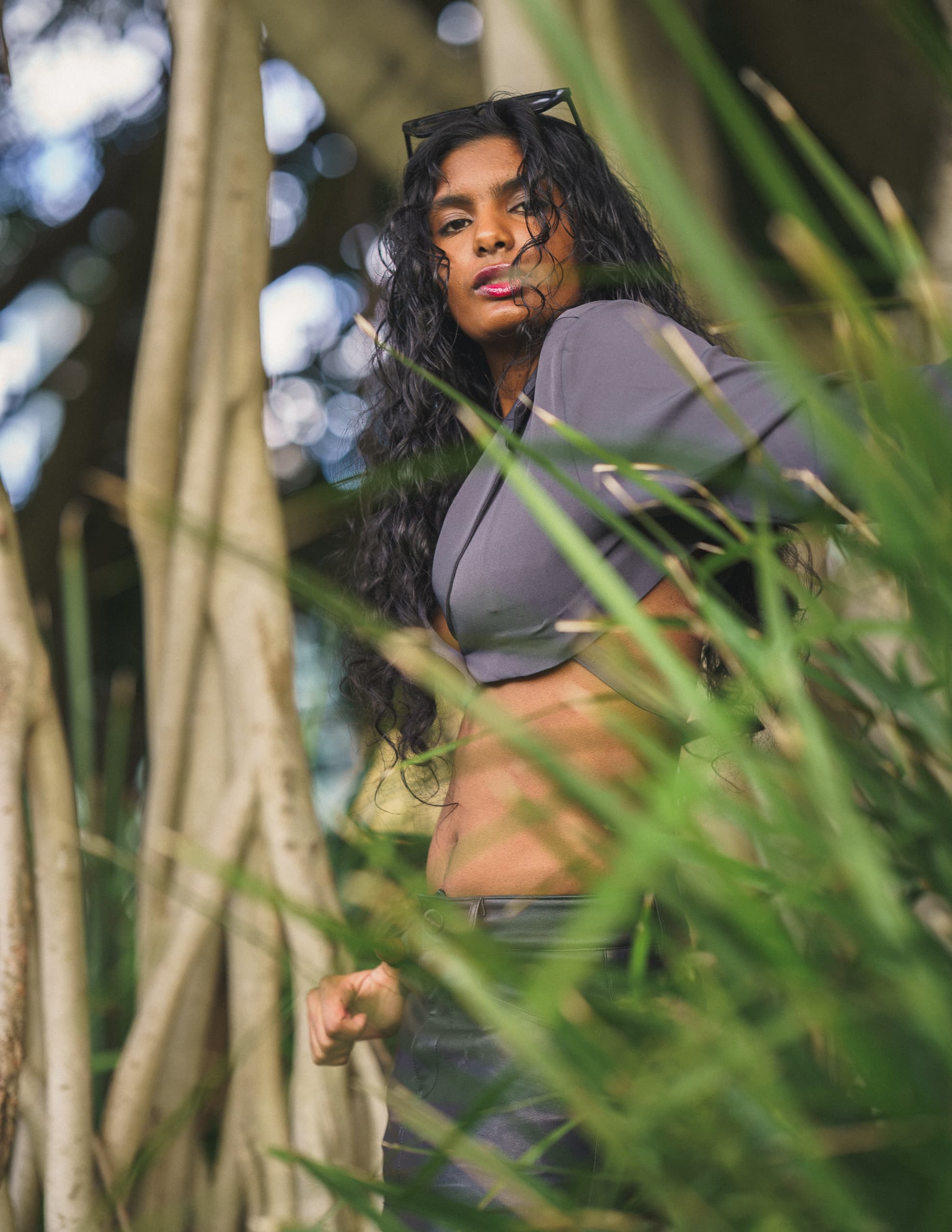 Woman in a grey crop top standing between banyan roots and tall blades of grass