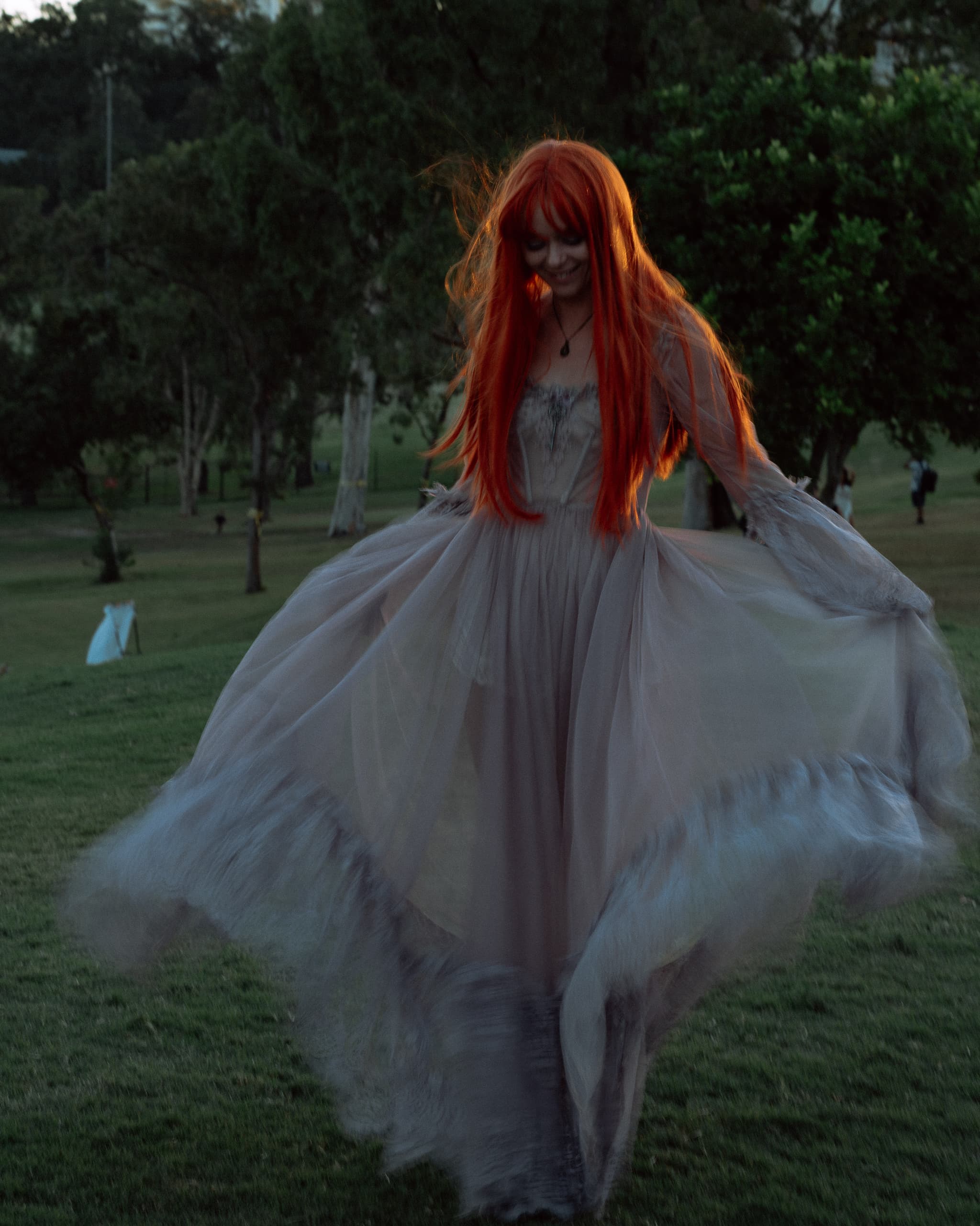 Woman with long red hair twirling a flowing pale dress on parkland at dusk