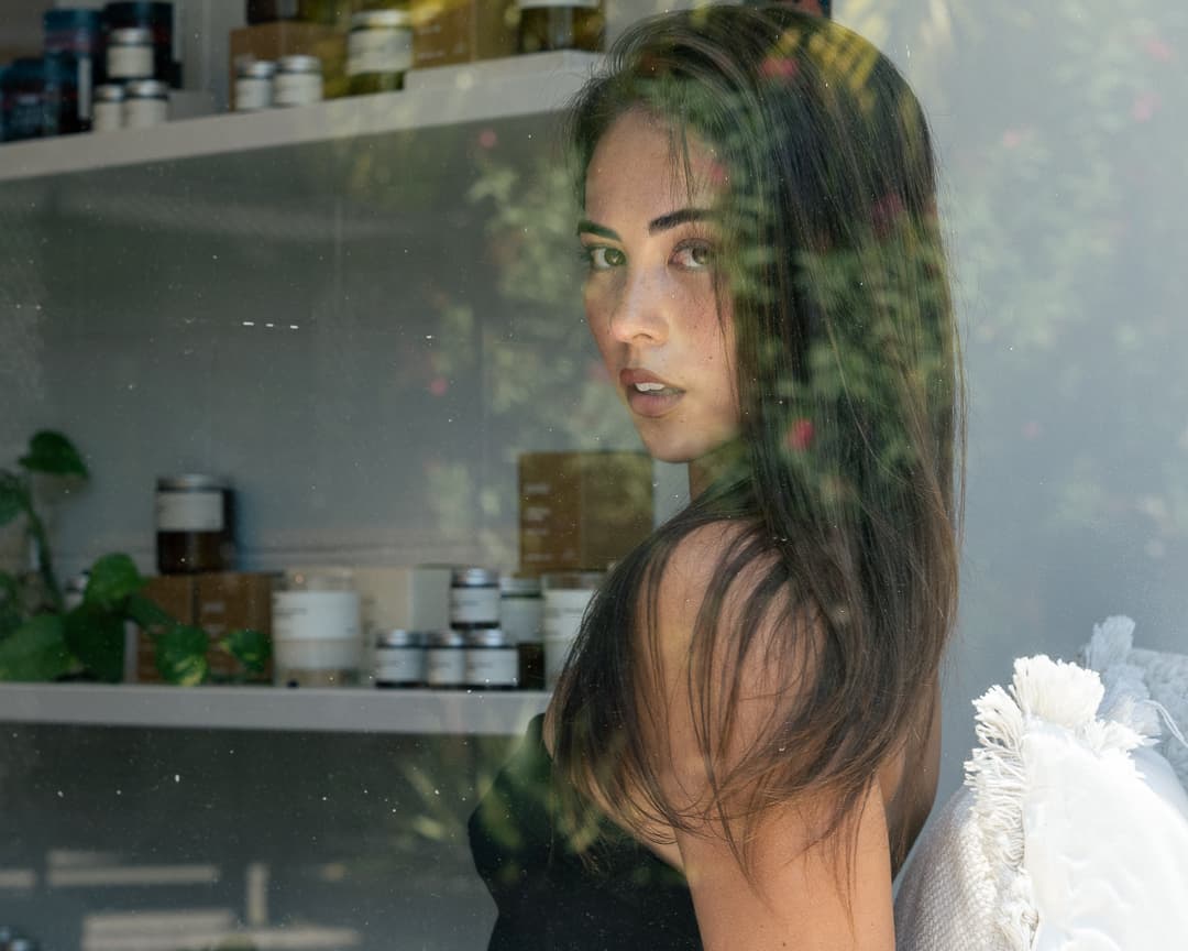 Woman glancing back through a shop window, shelves of jars reflected in the glass