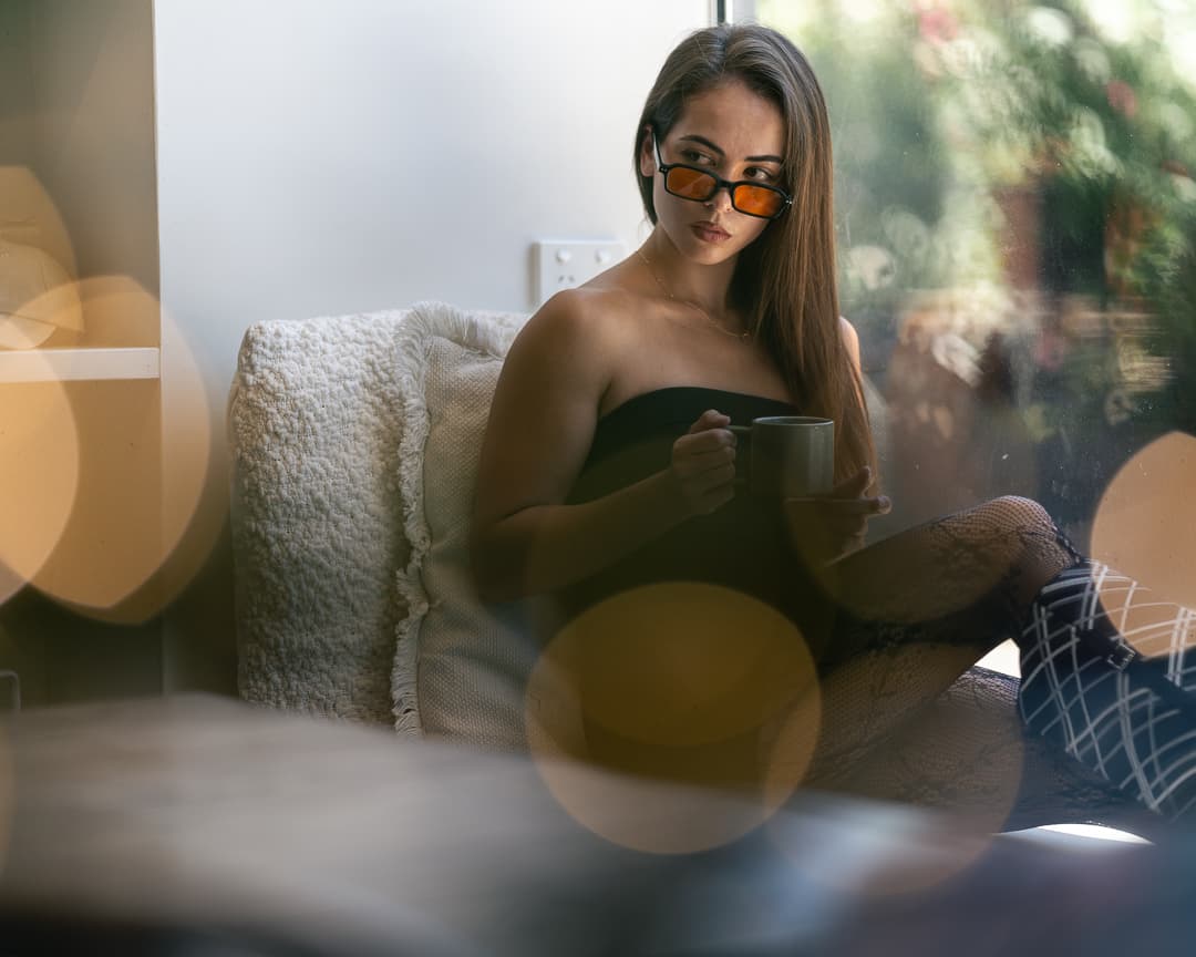 Woman in tinted sunglasses having coffee by a window, shot through warm bokeh