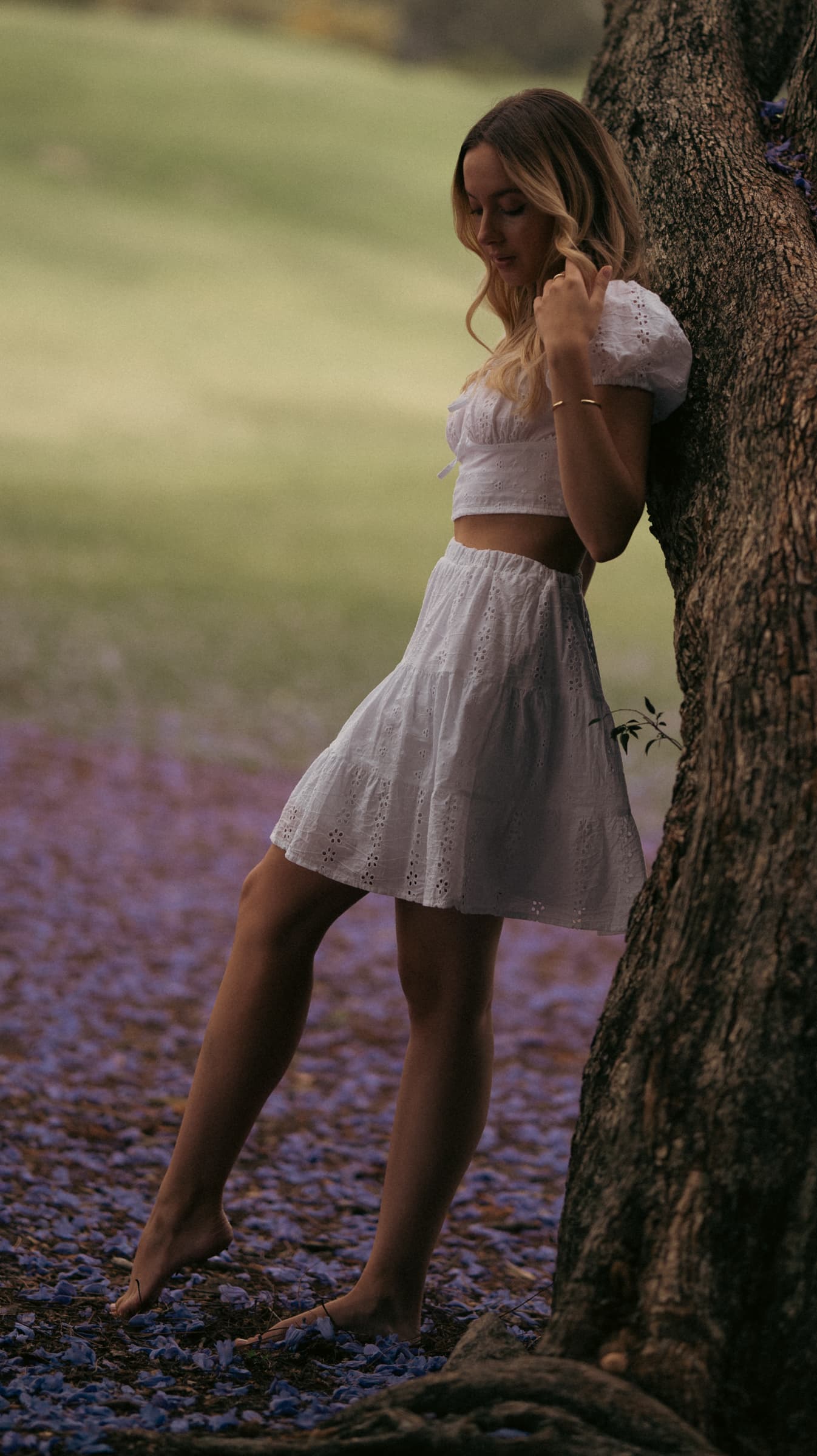 Woman in a white dress leaning against a tree trunk on a carpet of purple jacaranda flowers