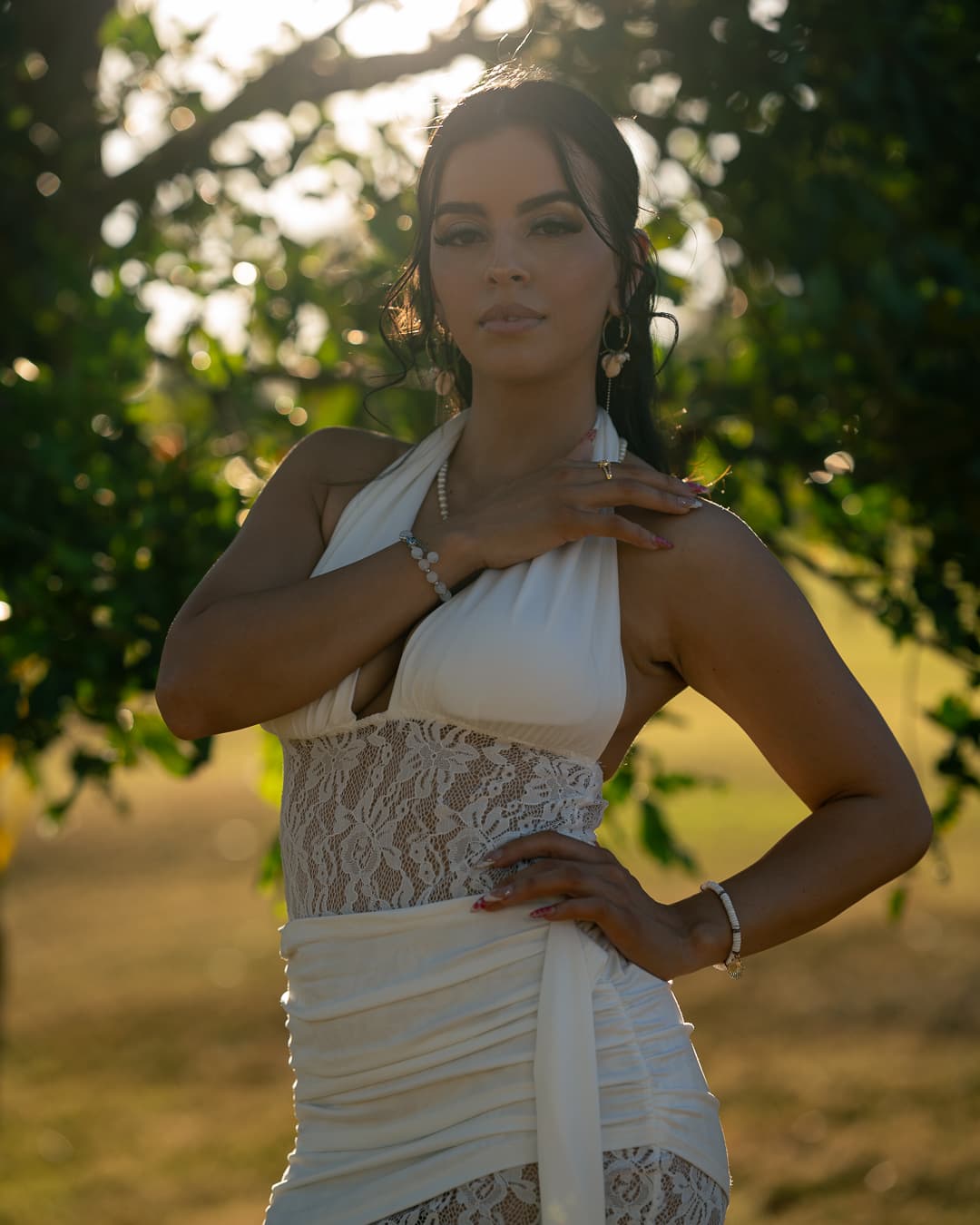Woman in a white lace halter dress under a tree, lit from behind by golden hour sun