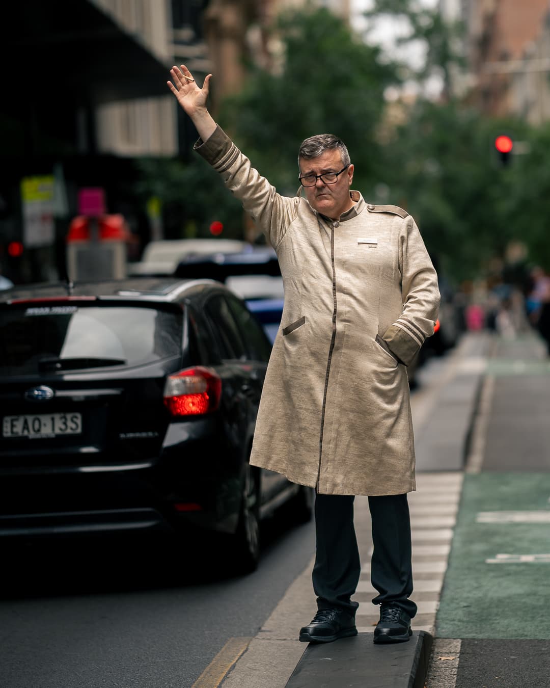 Man in a long beige coat hailing a cab with his arm raised high in city traffic