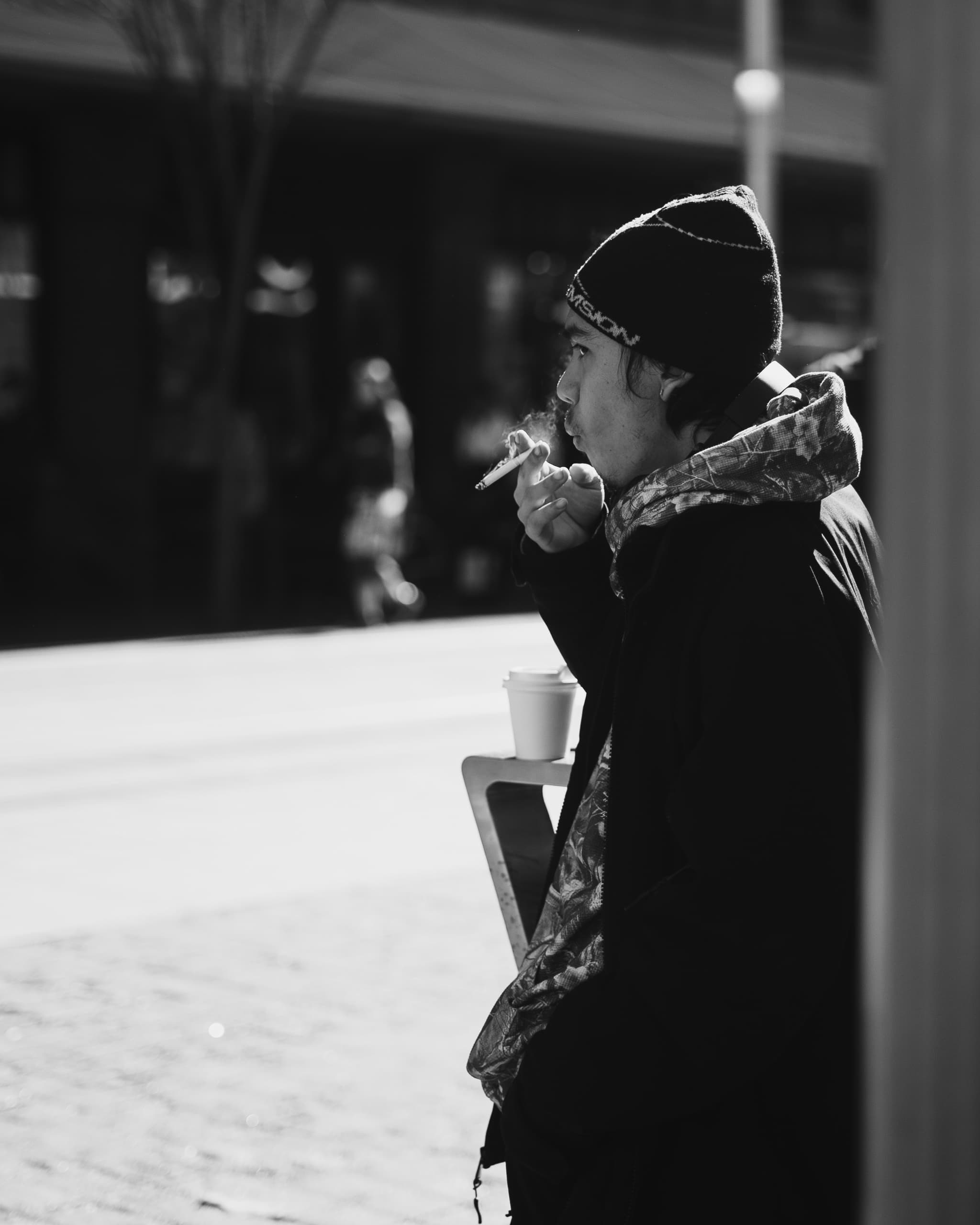 Black and white profile of a man in a beanie smoking over a takeaway coffee