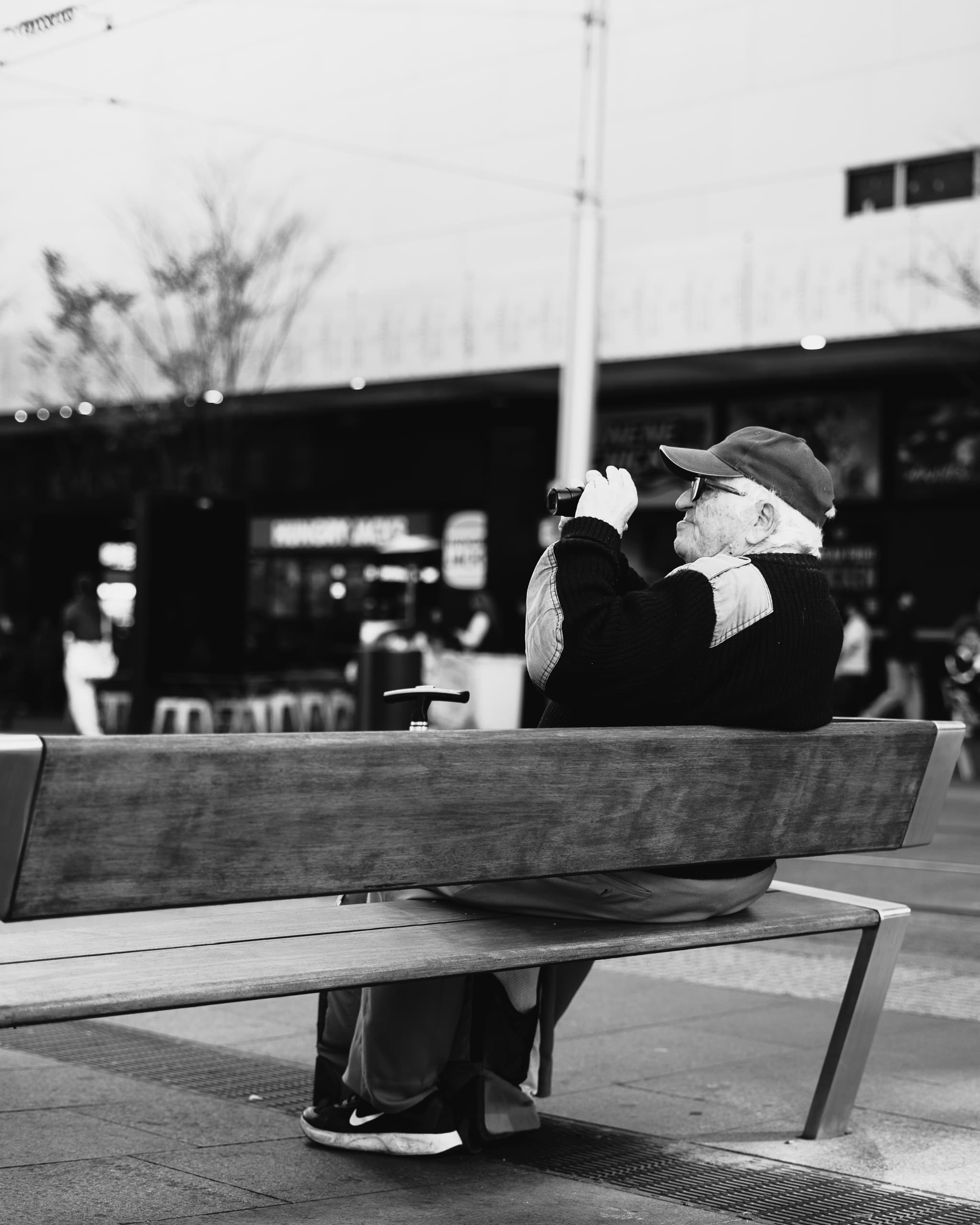 Black and white view of an older man on a bench scanning the square through binoculars