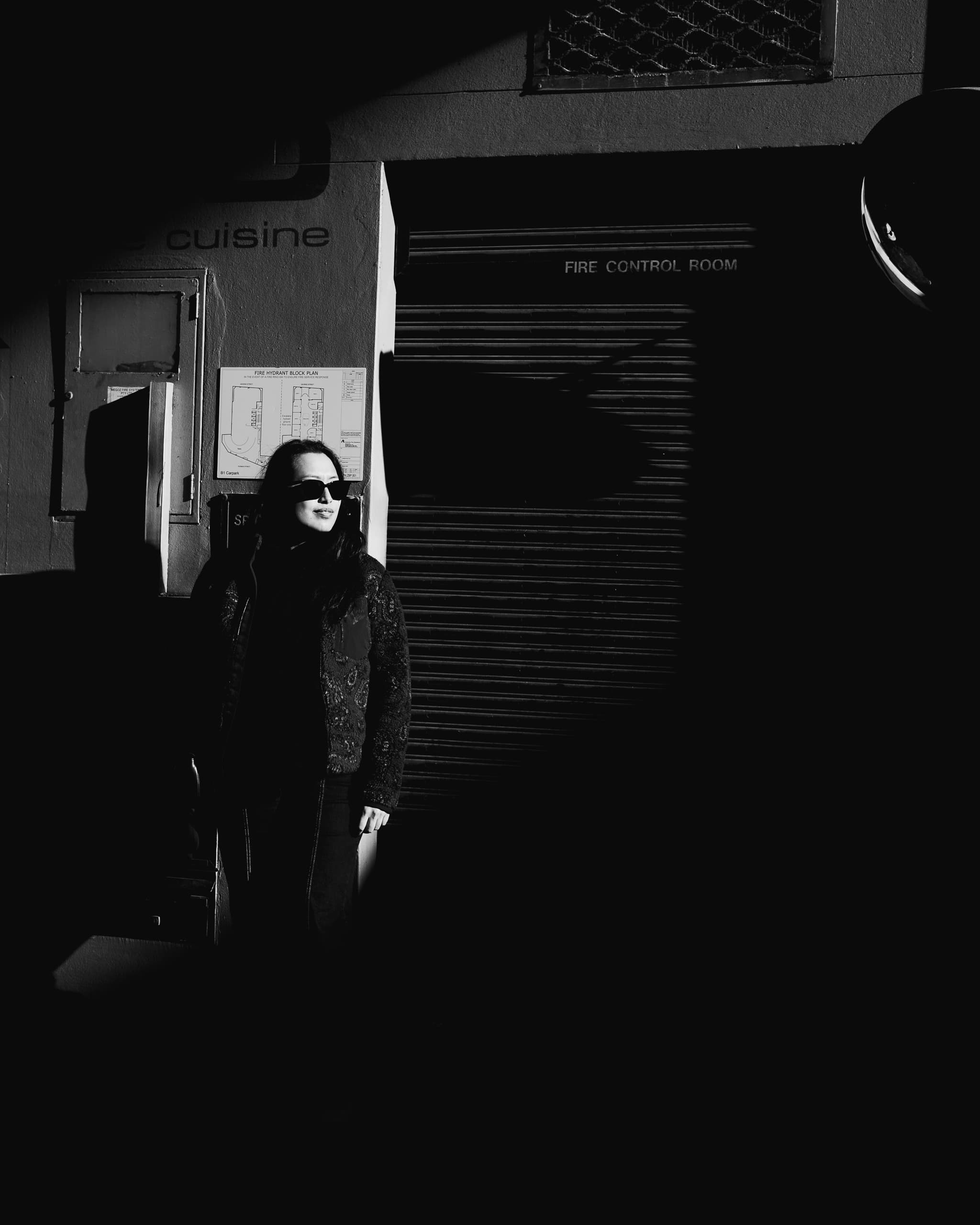 Black and white shot of a woman in sunglasses lit by a strip of sun on a dark wall