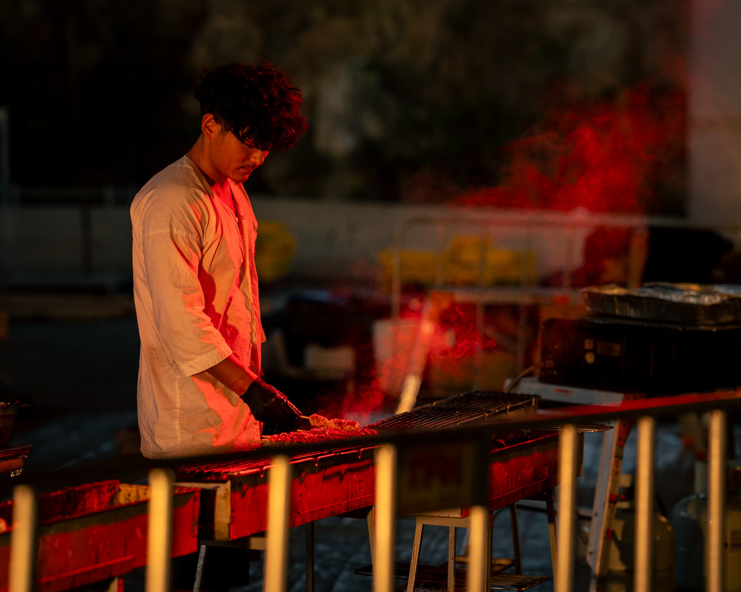 Cook in a white shirt grilling skewers behind a railing, bathed in red light