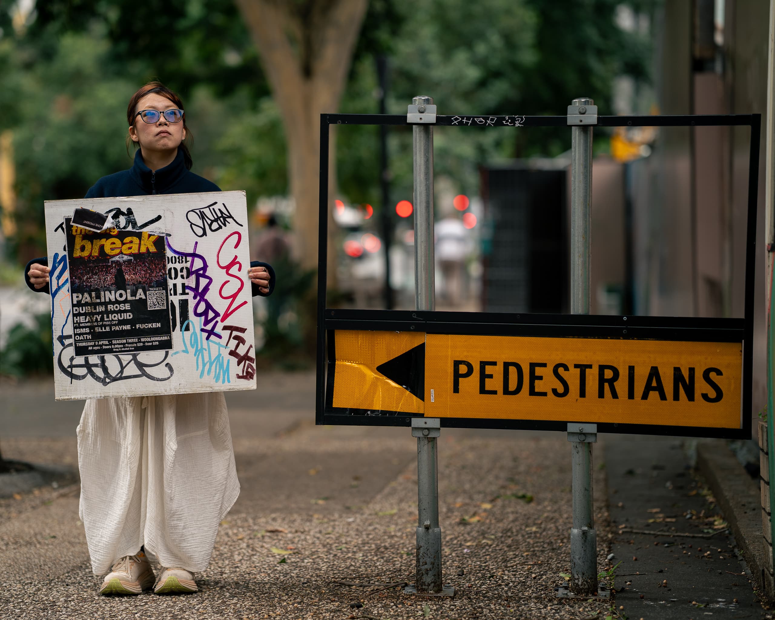 Woman holding a graffitied gig poster beside a yellow Pedestrians road sign