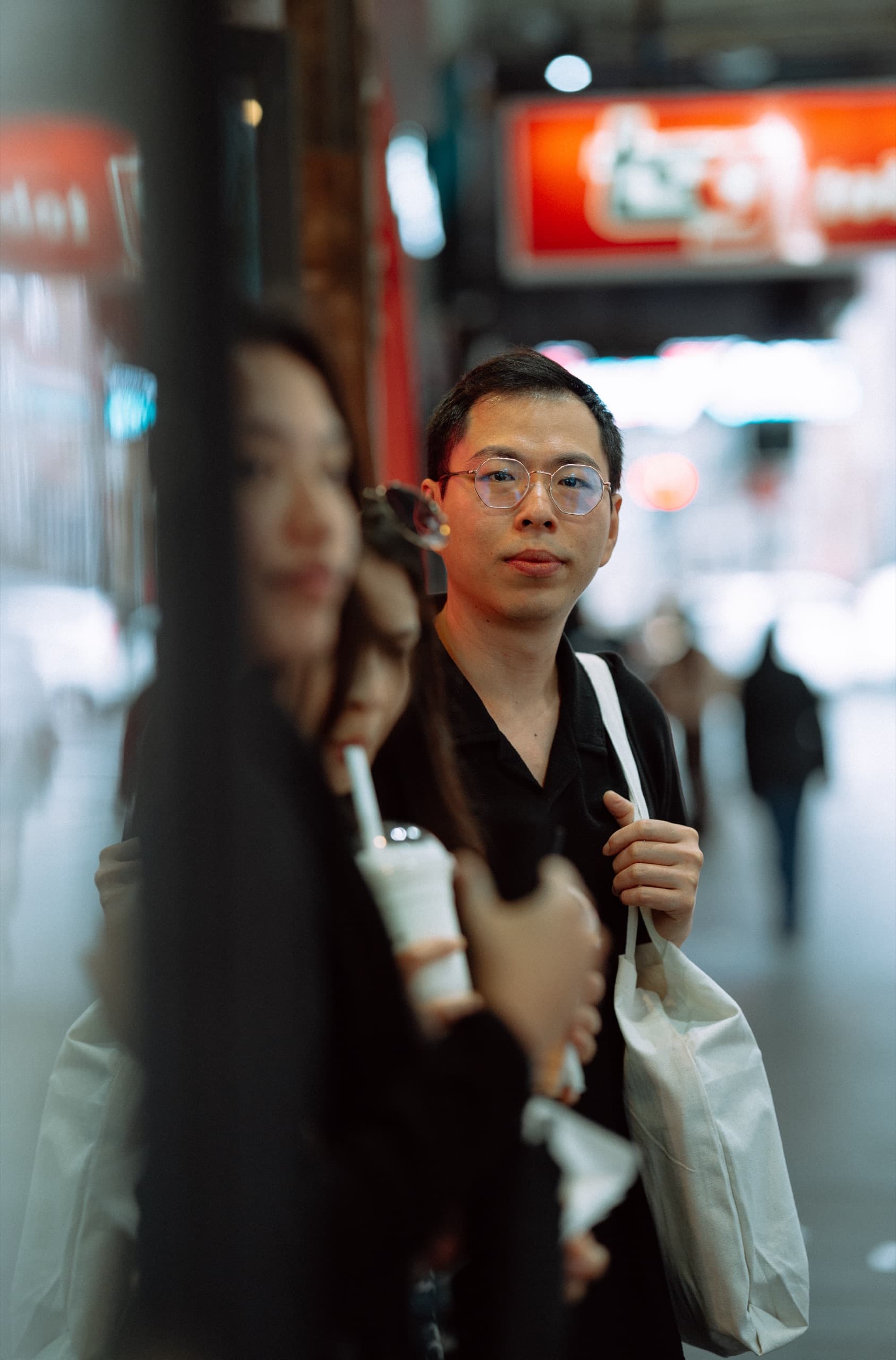 Man with glasses and a tote bag glancing at the camera on a neon-lit street at night