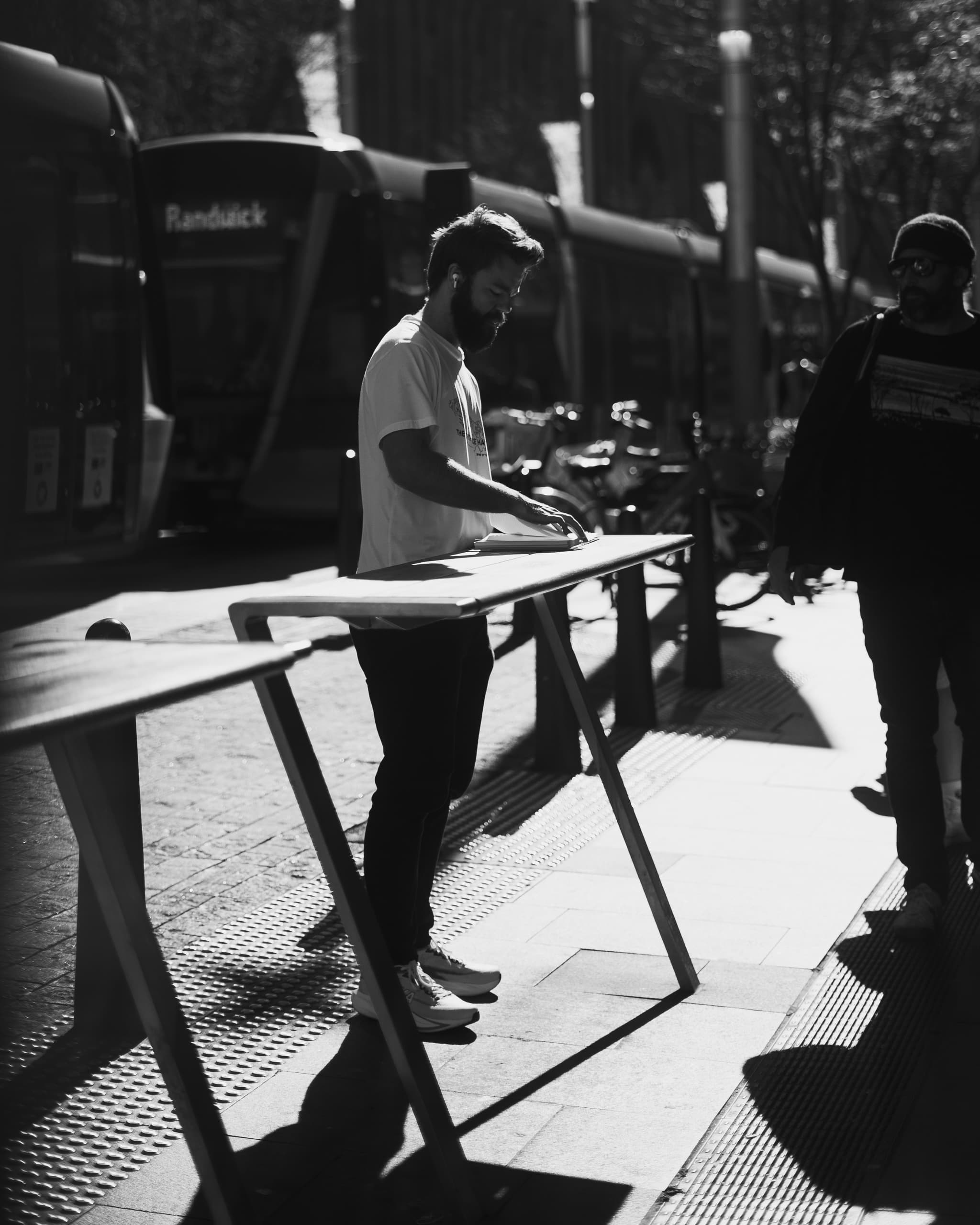 Black and white scene of a man writing at a standing table beside the light rail
