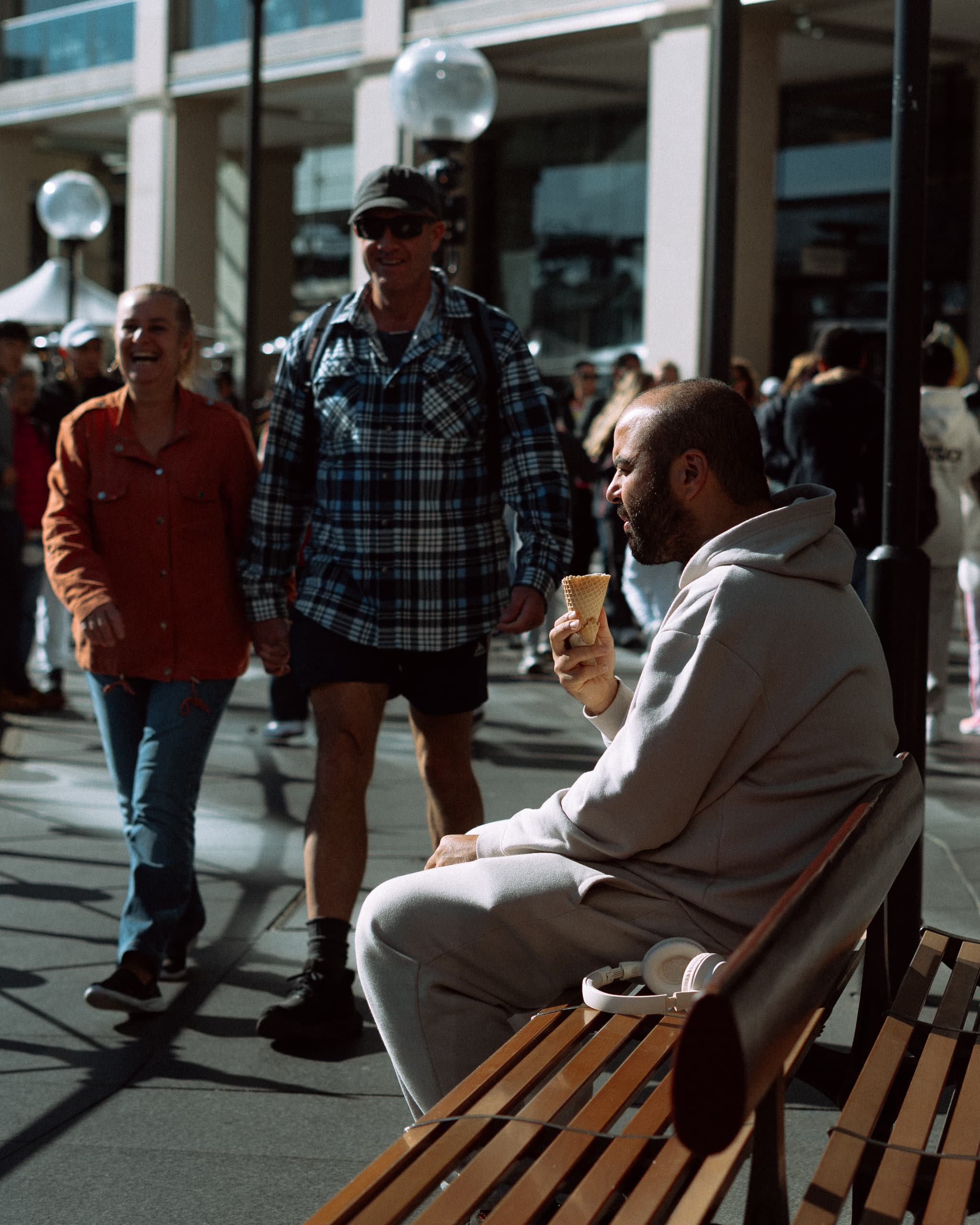 Man in a hooded tracksuit eating an ice cream cone on a bench in winter sun