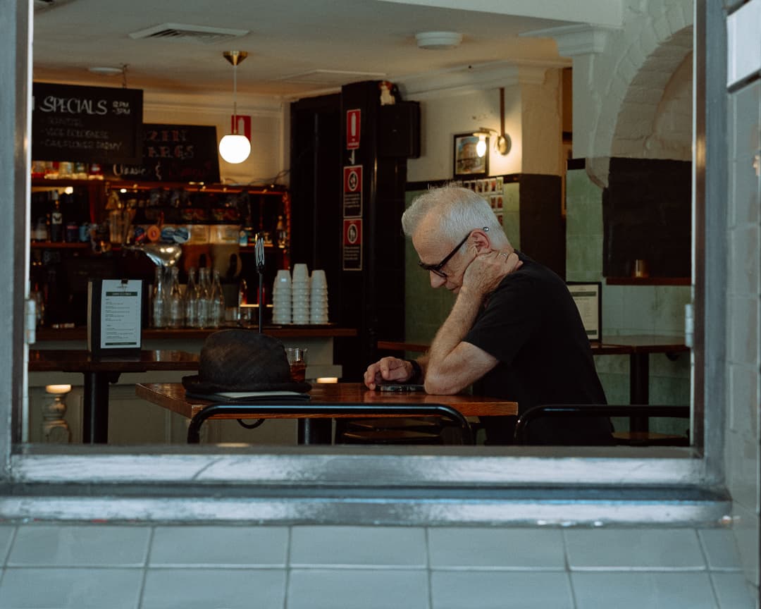 Older man sitting alone at a pub table, seen through the front window from the street