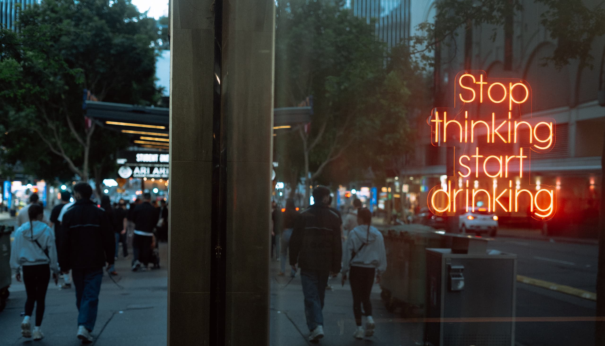 Neon window sign reading Stop thinking start drinking, evening crowd reflected in the glass