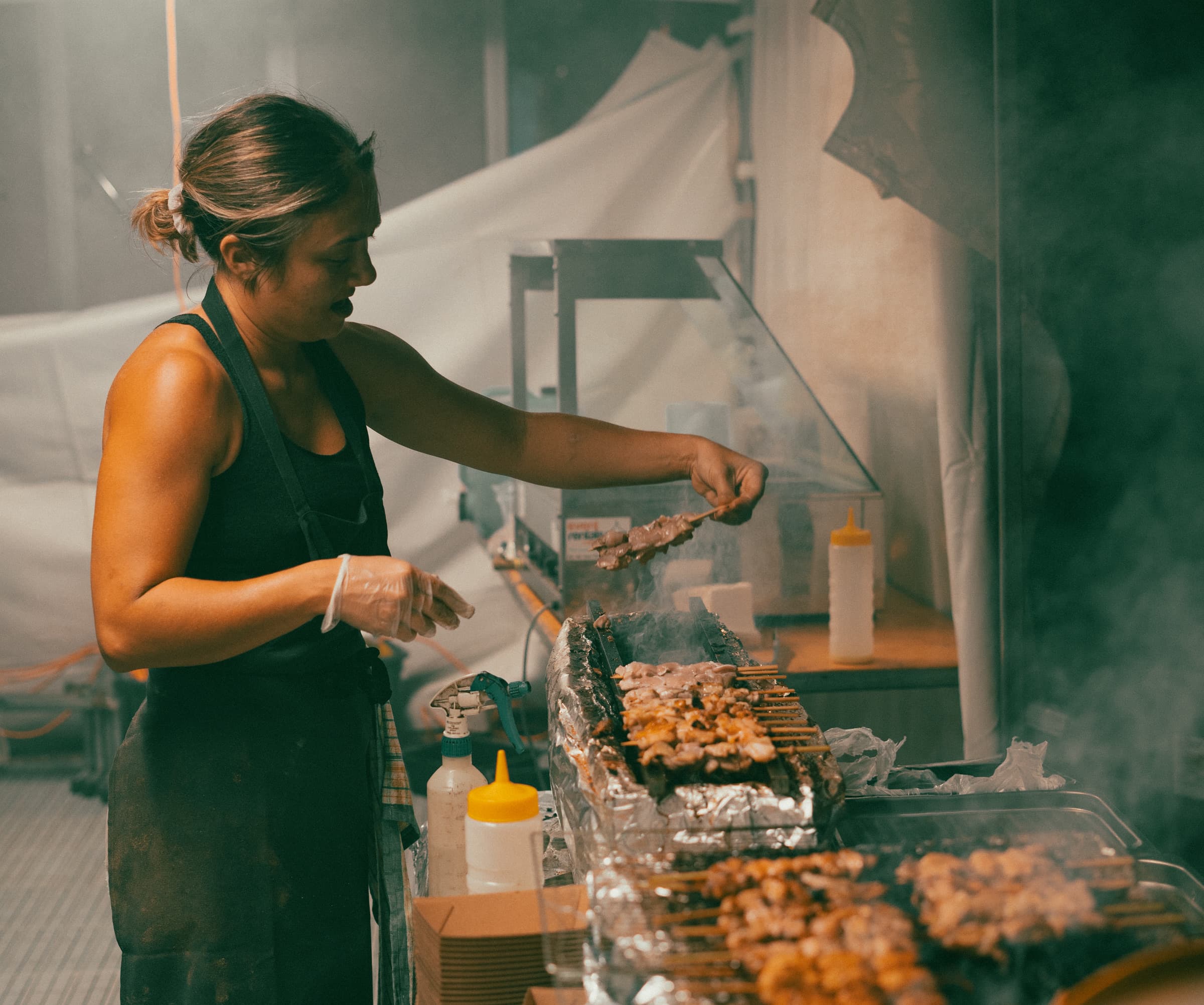 Street food cook turning skewers on a grill in a cloud of smoke at a night market