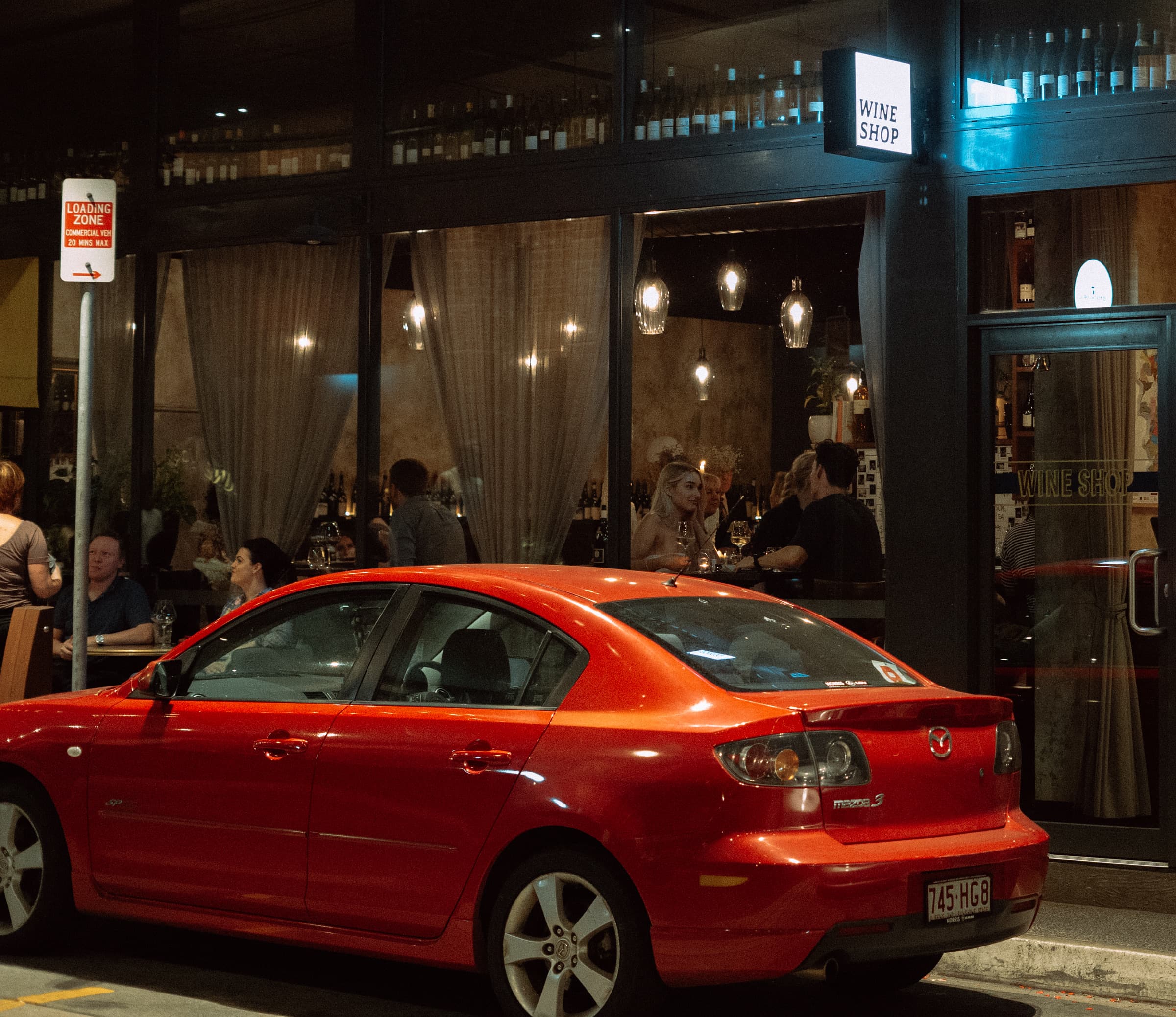 Red sedan parked outside a wine shop glowing with warm light at night