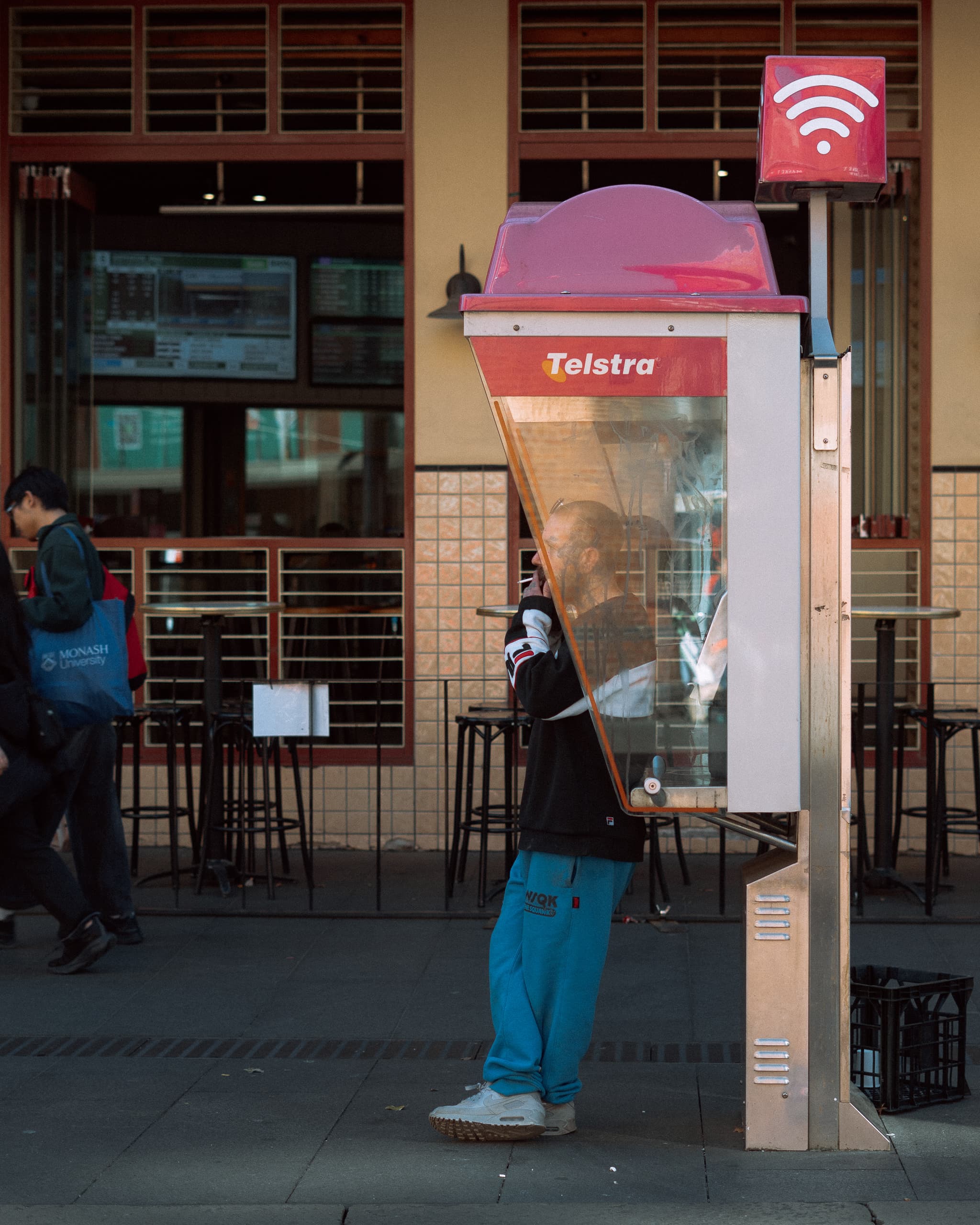 Man smoking beside a Telstra phone booth outside a tiled shopfront