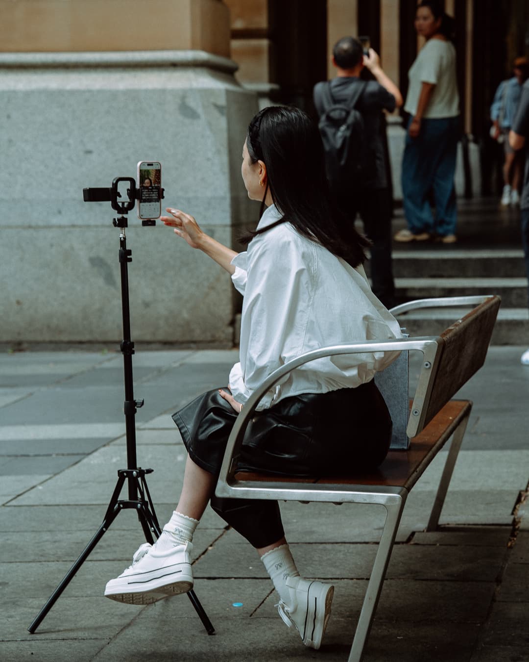 Young woman filming herself on a phone tripod while sitting on a city bench