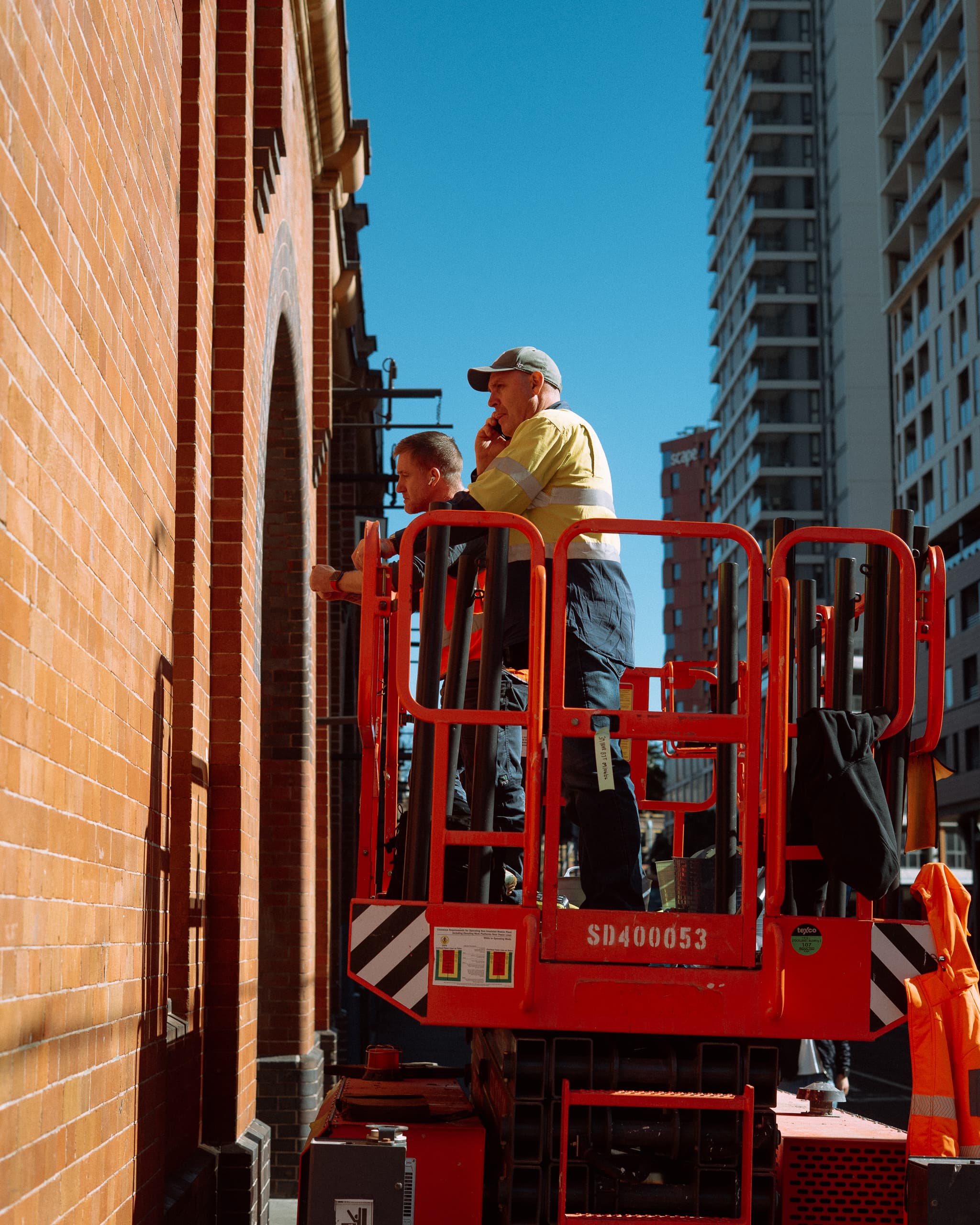Two workers on a scissor lift beside a red brick wall under deep blue sky